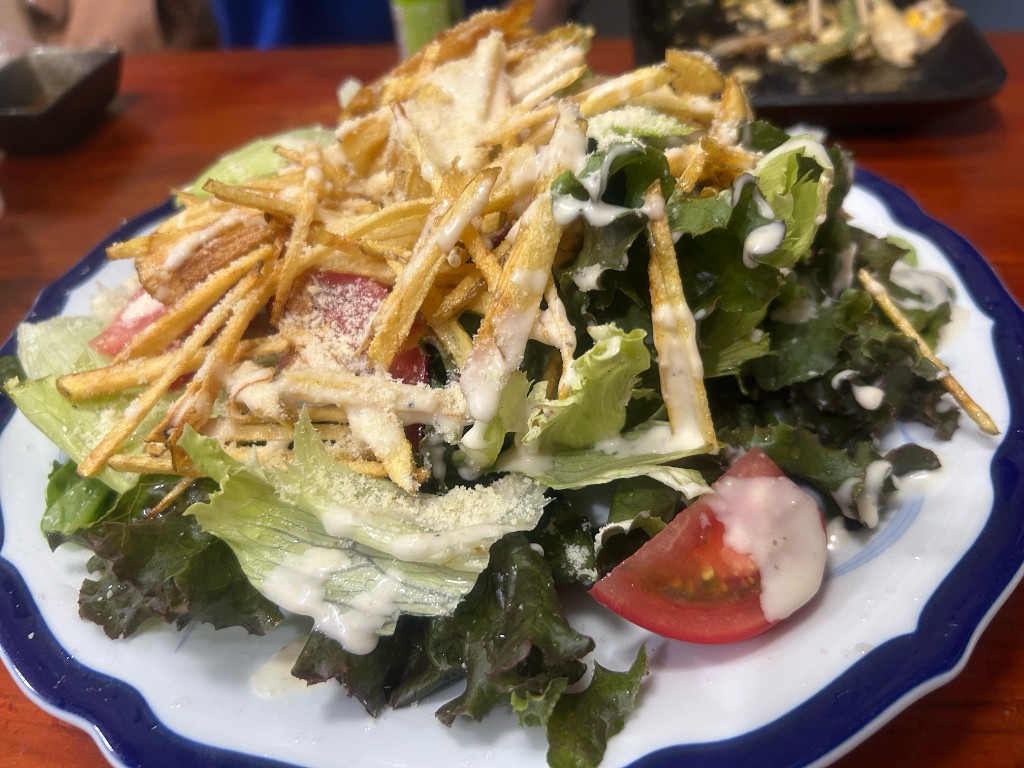白い縁取りのある皿に盛られた大きなサラダ。細く揚げた芋(またはパスタ)が山盛りに載り、ドレッシングがかかっている(居酒屋ふる里 麻生店)