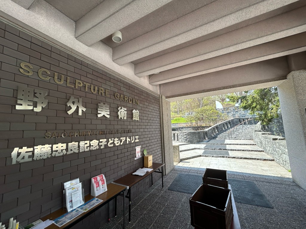 「野外美術館」「佐藤忠良記念子どもアトリエ」の看板が並ぶ入口。パンフレットラック越しに石段と緑が見える（札幌芸術の森）