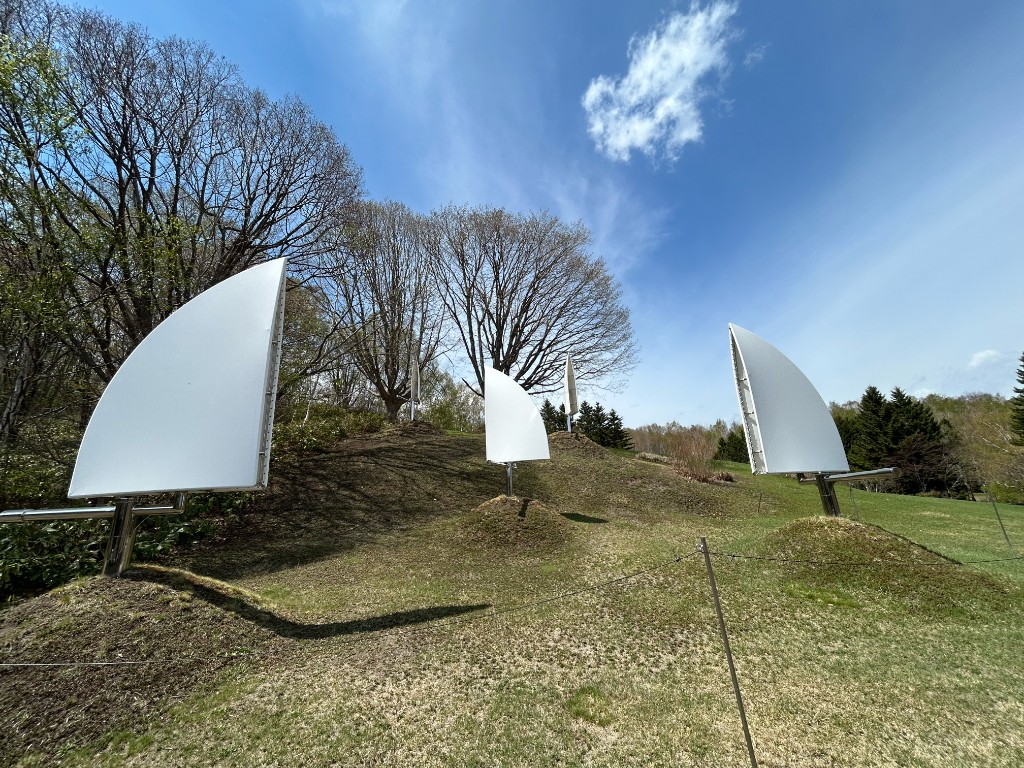 なだらかな丘の上に、白い帆のような大型彫刻が複数配置されている。青空と新緑が広がる（新宮晋「雲の牧場」・札幌芸術の森）