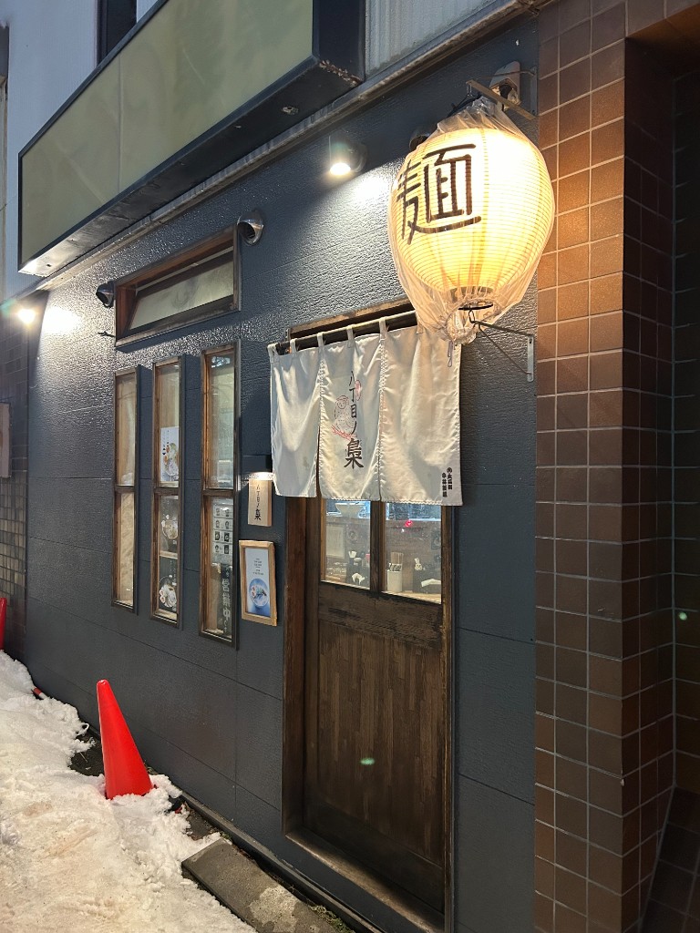 夜の外観。「麺」の提灯と暖簾、雪の残る街角の雰囲気