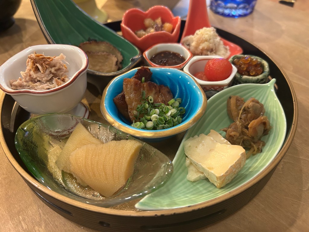 黒い丸い盆に並んだ八寸。だいこんの煮物、鹿肉の煮物、とろろ、いくら、トマト、チーズなど多彩な小鉢が円形に配置される(酒家 ハル。)