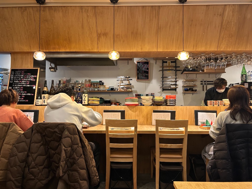 カウンター席に座る客と、黒板メニューを前に立つ店主。木のカウンターと温かい照明、背後に並ぶ日本酒の瓶(酒家 ハル。)