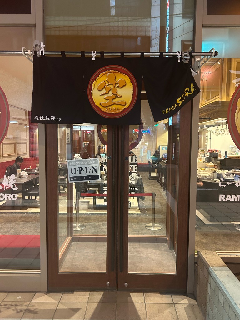 黒地に金の「空」マークが入った暖簾。「RAMEN SORA」「森住製麺より」の文字。OPEN の札が下がる店内入口(らーめん空)